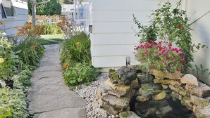Un chemin de pierres mène à une maison qui possède un petit bassin en pierre, ainsi que diverses plantes et fleurs.