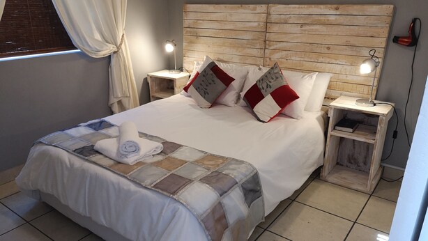 Cama bem arrumada com colcha, travesseiros e uma toalha enrolada. Duas mesinhas de cabeceira com luminárias, uma cabeceira de madeira e uma janela com cortinas.