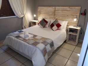 A neatly made bed with a quilt, pillows, and a rolled towel. There are two bedside tables with lamps, a wooden headboard, and a window with curtains. - Smithland Guest Apartments (Cape Town)