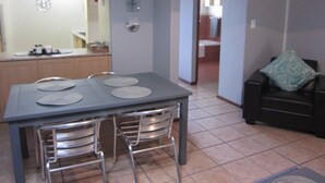 Uma sala de jantar com mesa e cadeiras, uma cozinha ao fundo e uma sala de estar com sofá e almofada.