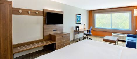 A hotel room with a large bed, a desk with a chair, a flat-screen TV, a small table, and a window with a view of trees.
