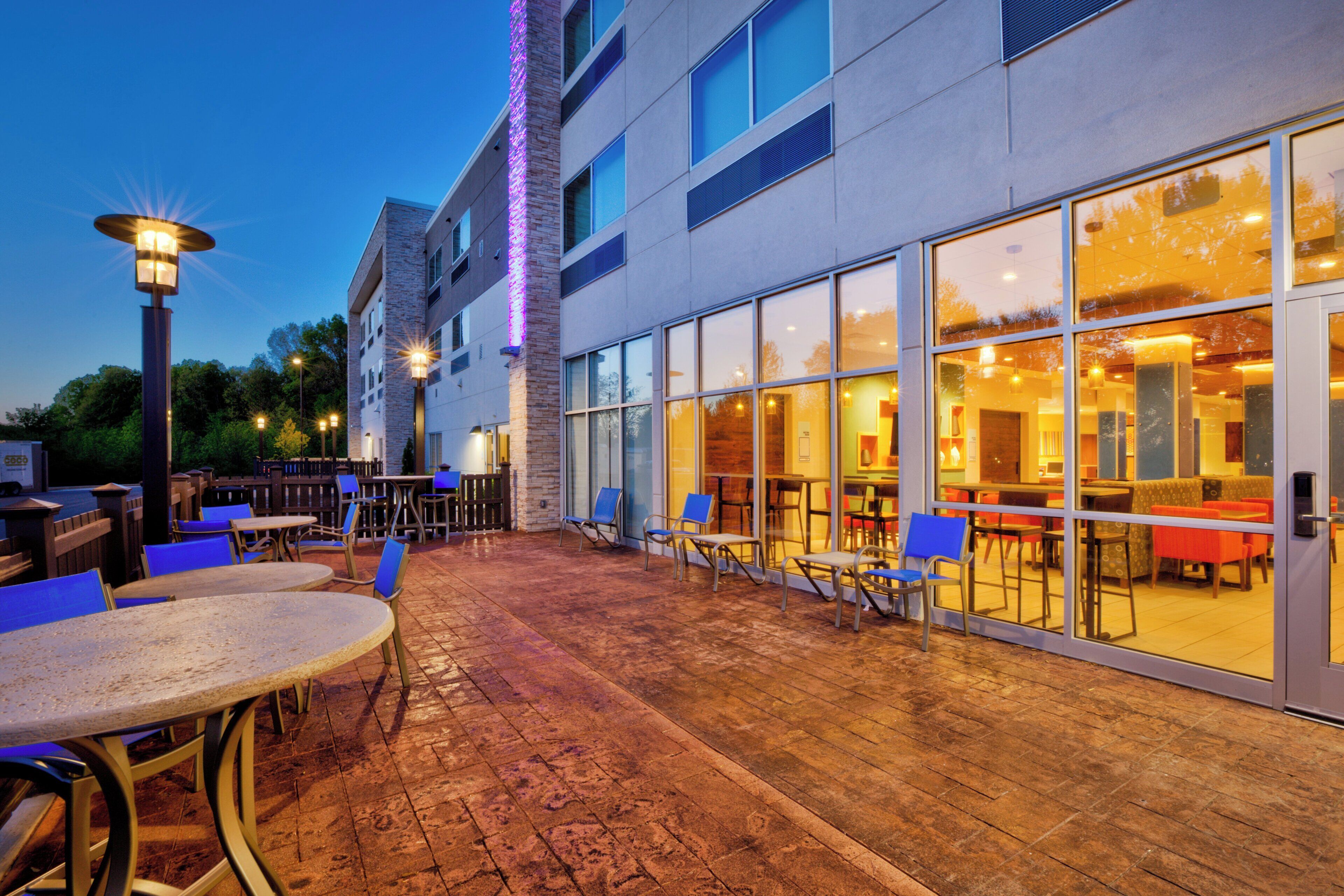 An outdoor patio area with tables and chairs, a brick floor, and a building with large windows and a stone facade.