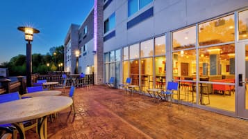 An outdoor patio area with tables and chairs, a brick floor, and a building with large windows and a stone facade.