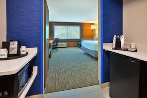 Quarto de hotel com cama, escrivaninha, micro-ondas, geladeira e janela com vista para árvores.