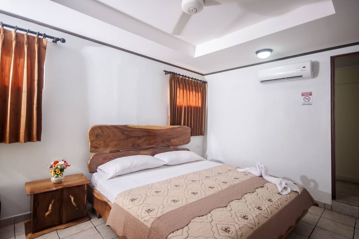 Une chambre avec un lit en bois, du linge de lit blanc, une table de chevet ornée d’un vase de fleurs et un appareil de climatisation fixé au mur.