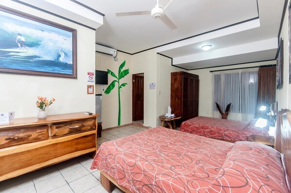 Habitación de hotel con dos camas, un aparador de madera, ventilador de techo, una ventana grande con cortinas y televisor montado en la pared.