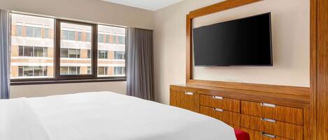 A hotel room with a large bed, a flat-screen TV mounted on the wall, a wooden dresser, and a window with curtains.