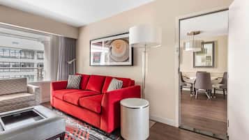 A modern hotel room with a red sofa, a grey armchair, a standing lamp, and a dining area visible through an open door.