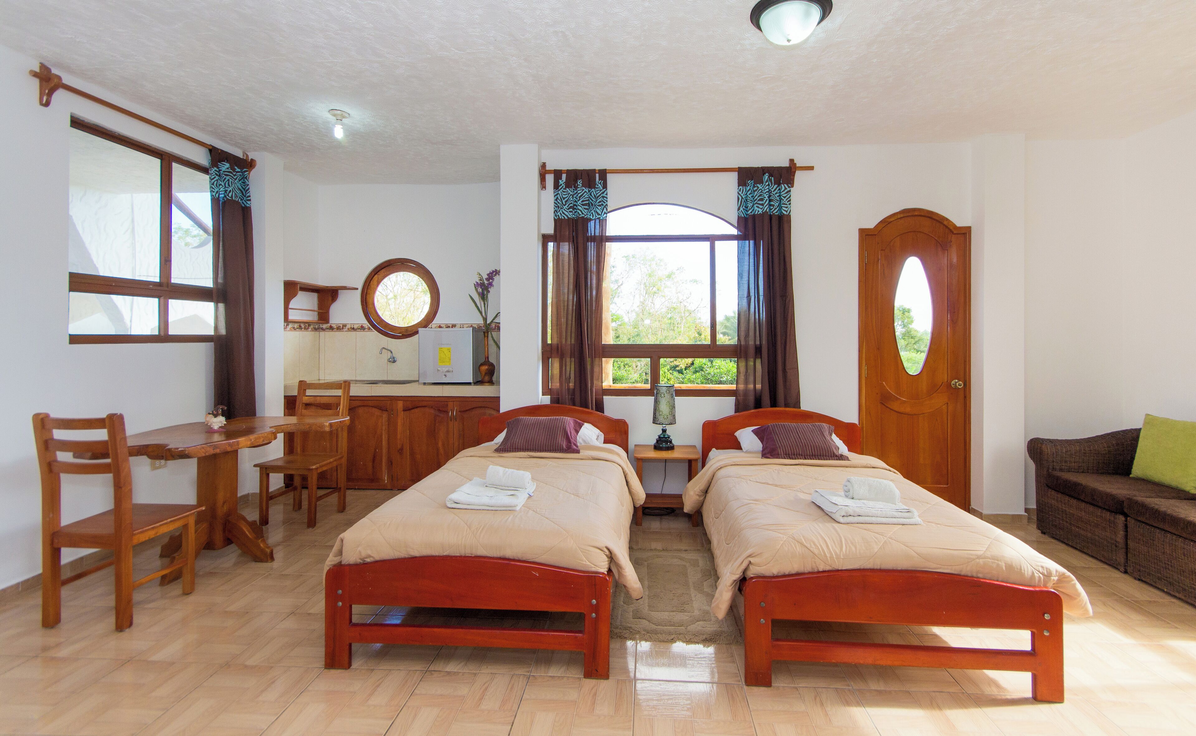 A room with two wooden beds, a wooden desk, a chair, a small table, a window with curtains, and a door.