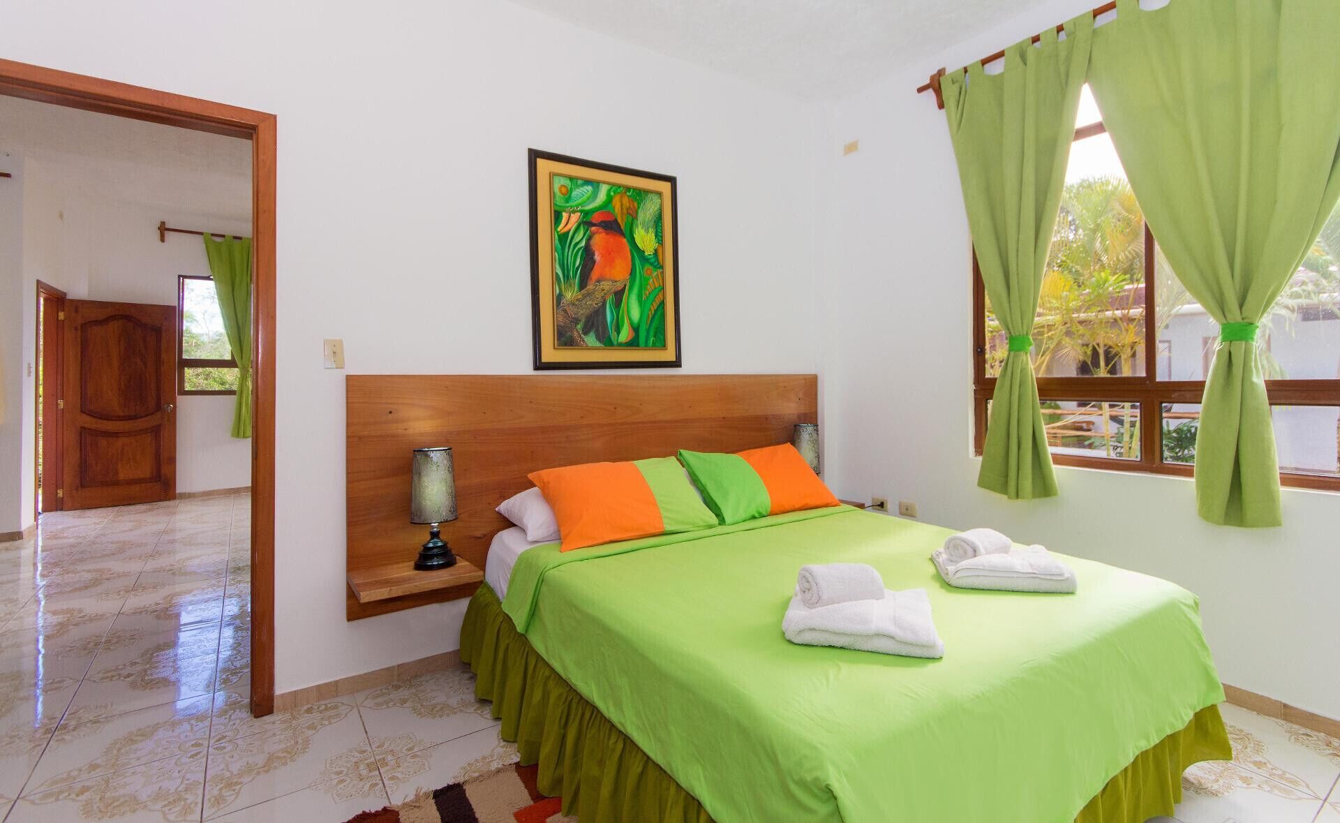 Quarto de hotel com cama, mesa de cabeceira, luminária, pintura e janela com cortinas.