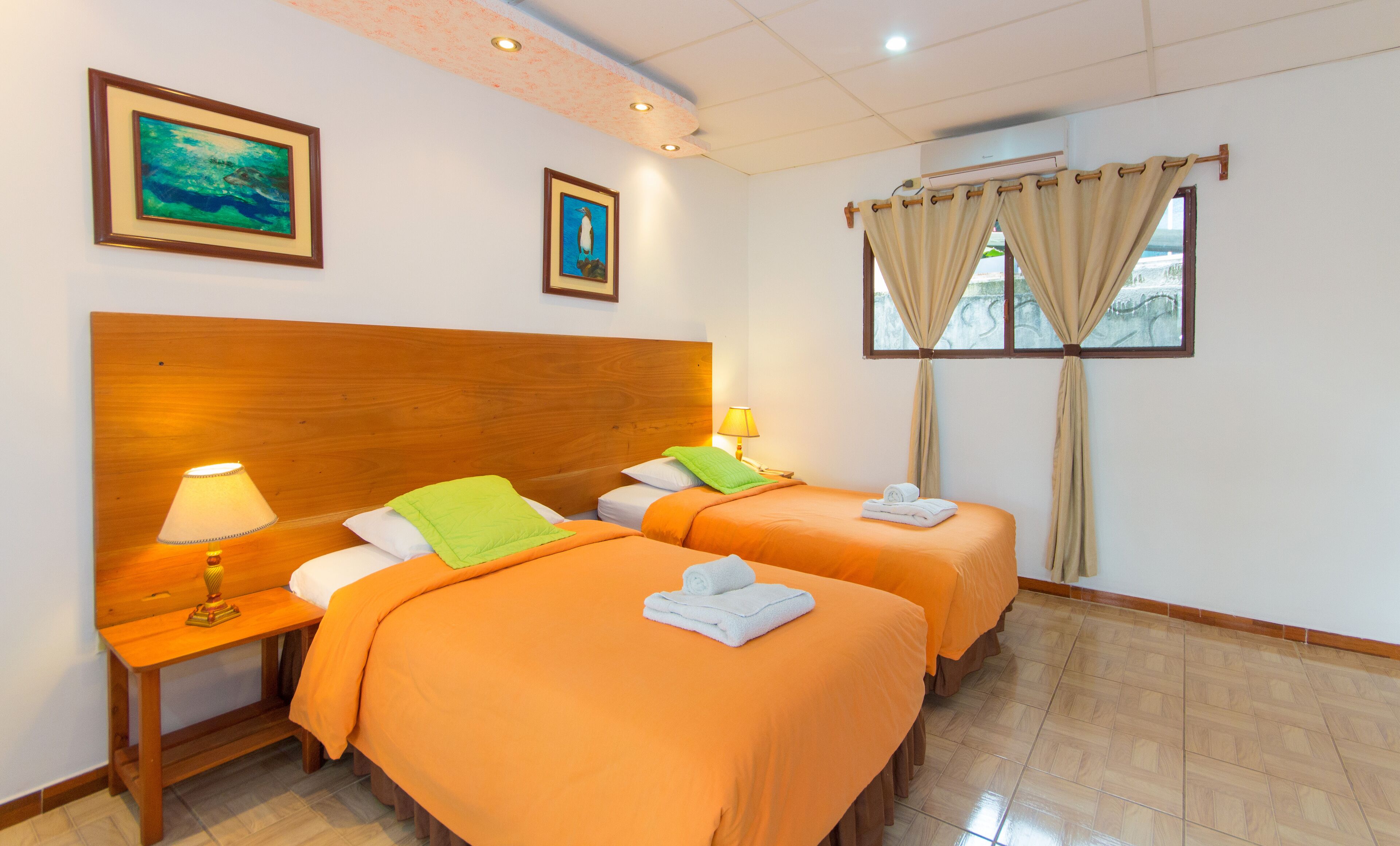 A hotel room with two beds, wooden headboards, a nightstand with a lamp, and a window with curtains.