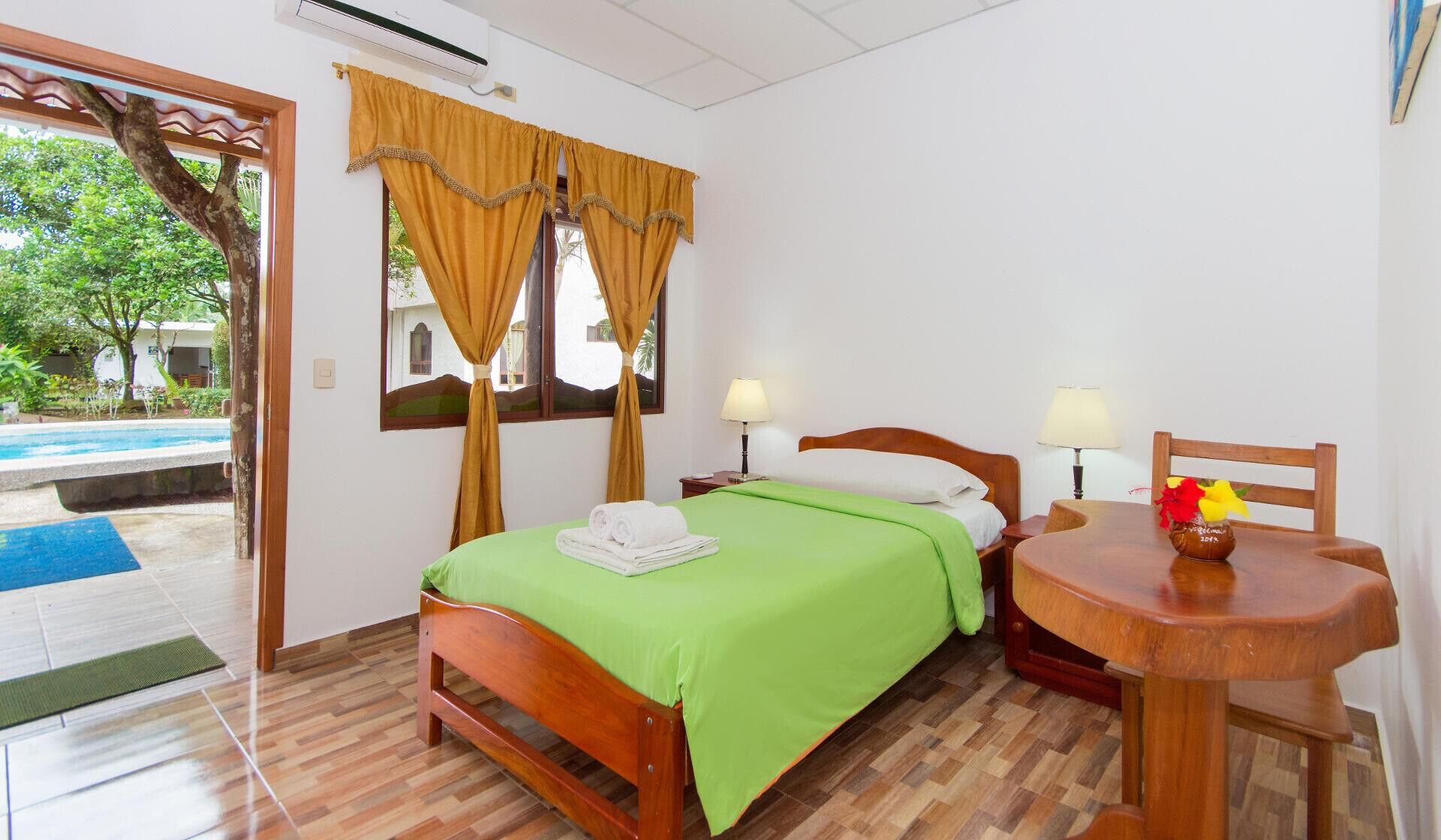 Quarto de hotel com uma cama, duas mesas de cabeceira com abajures, uma cadeira de madeira, uma pequena mesa com um vaso, e vista para uma piscina e vegetação lá fora.