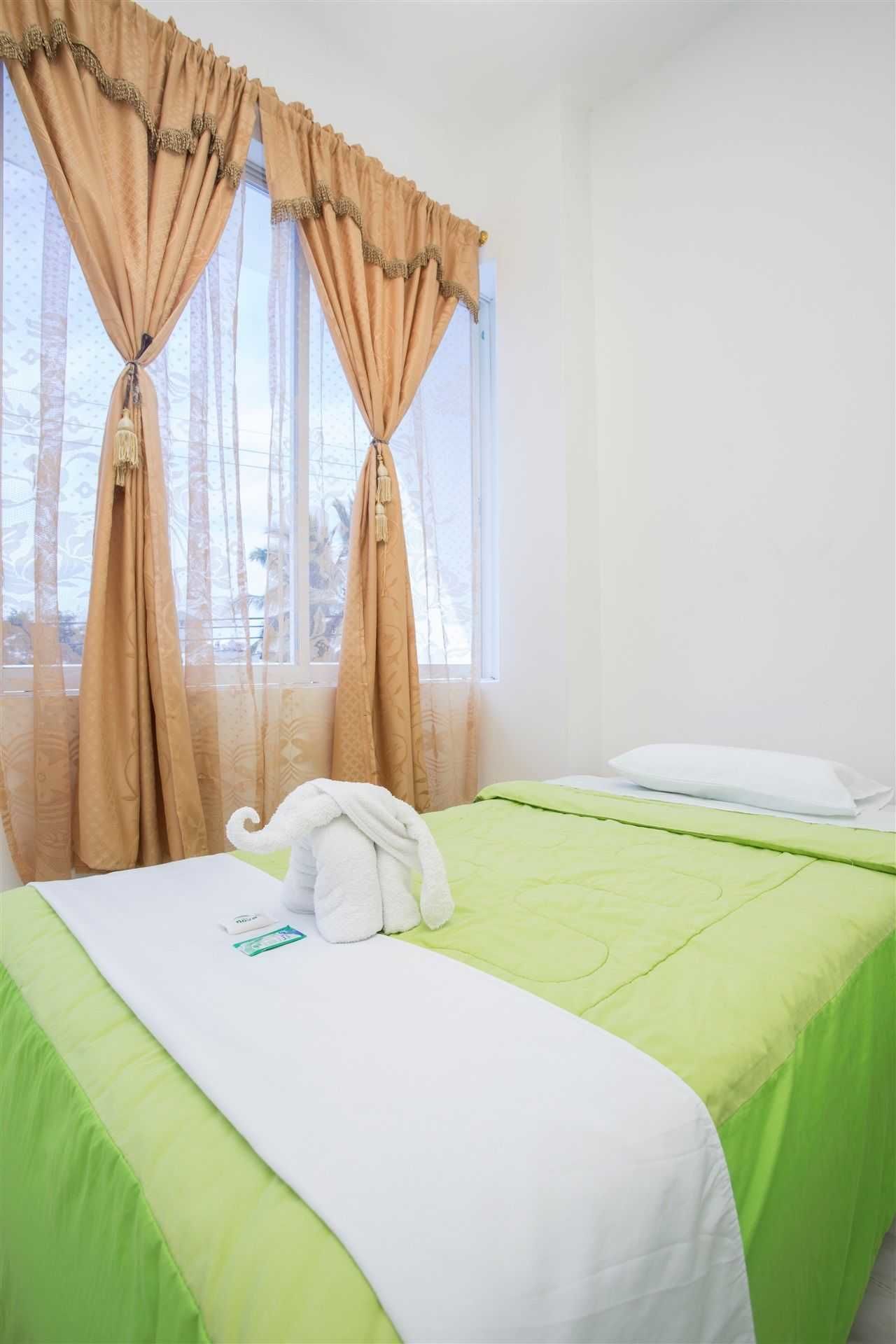 A hotel room with a bed, green bedding, a white pillow, and beige curtains.