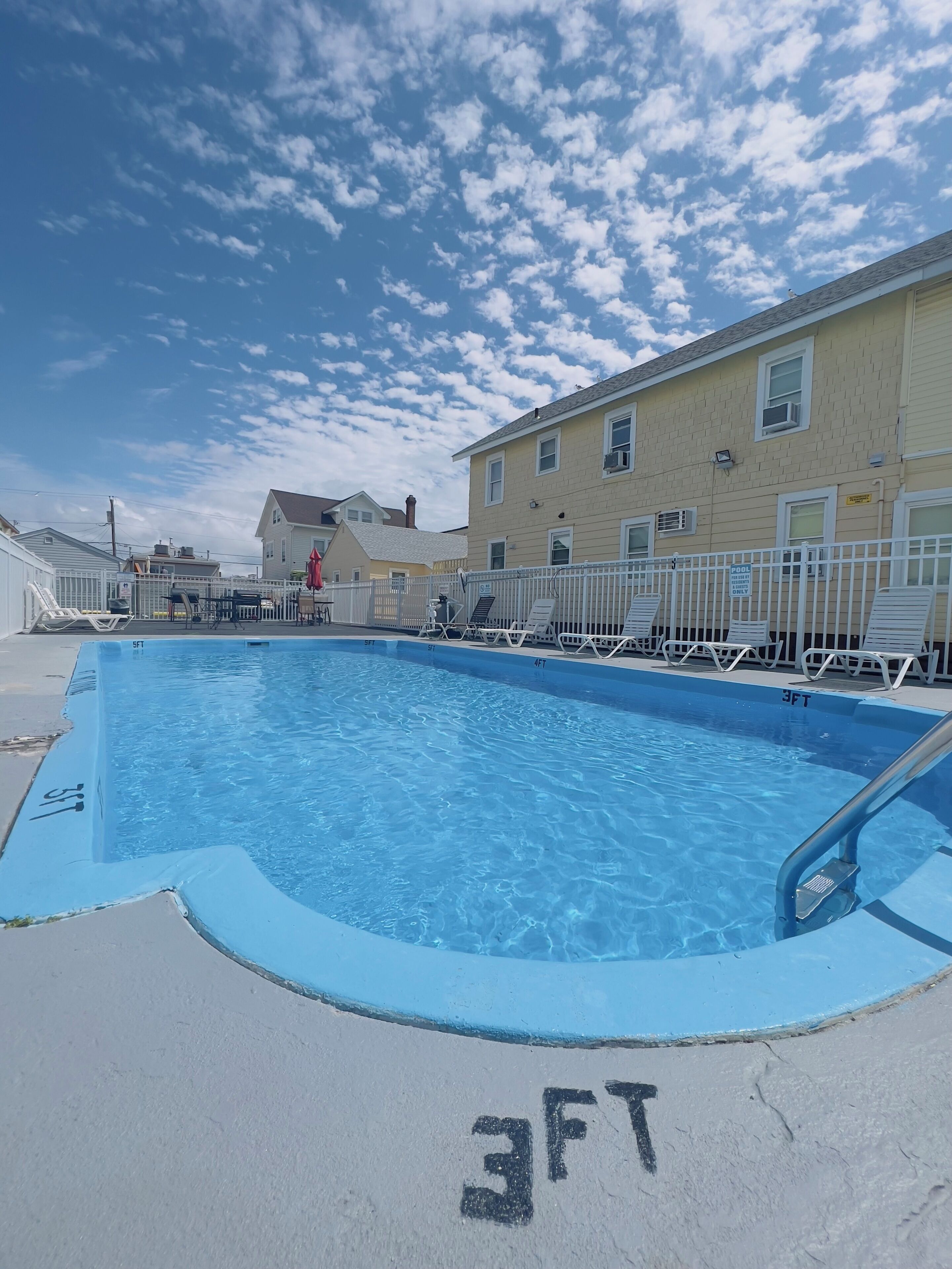 A rectangular pool with a 3FT depth marking, surrounded by a white fence and lounge chairs.