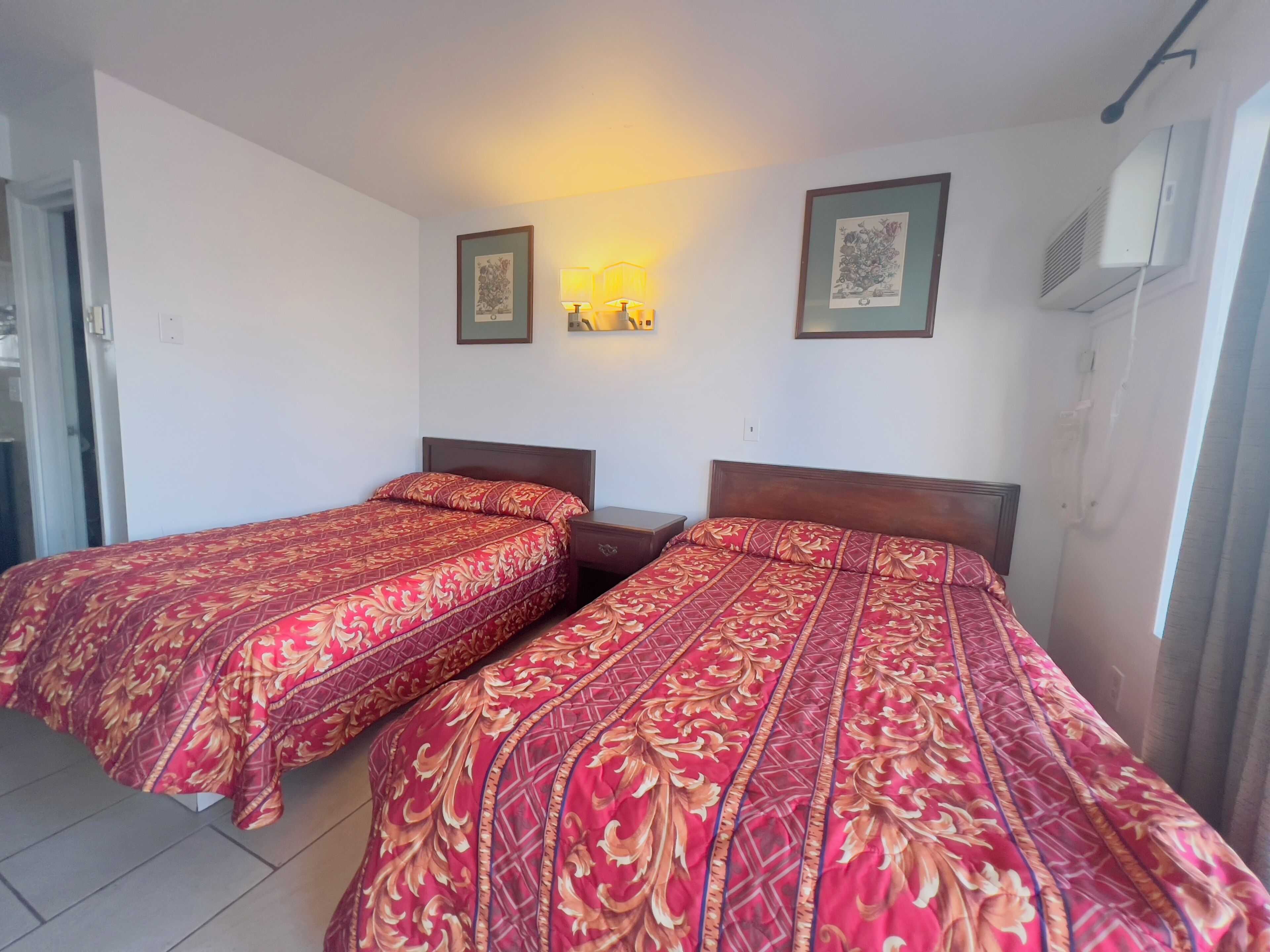 A hotel room with two beds, each with a patterned bedspread, a nightstand, a wall-mounted lamp, and framed artwork above the beds.