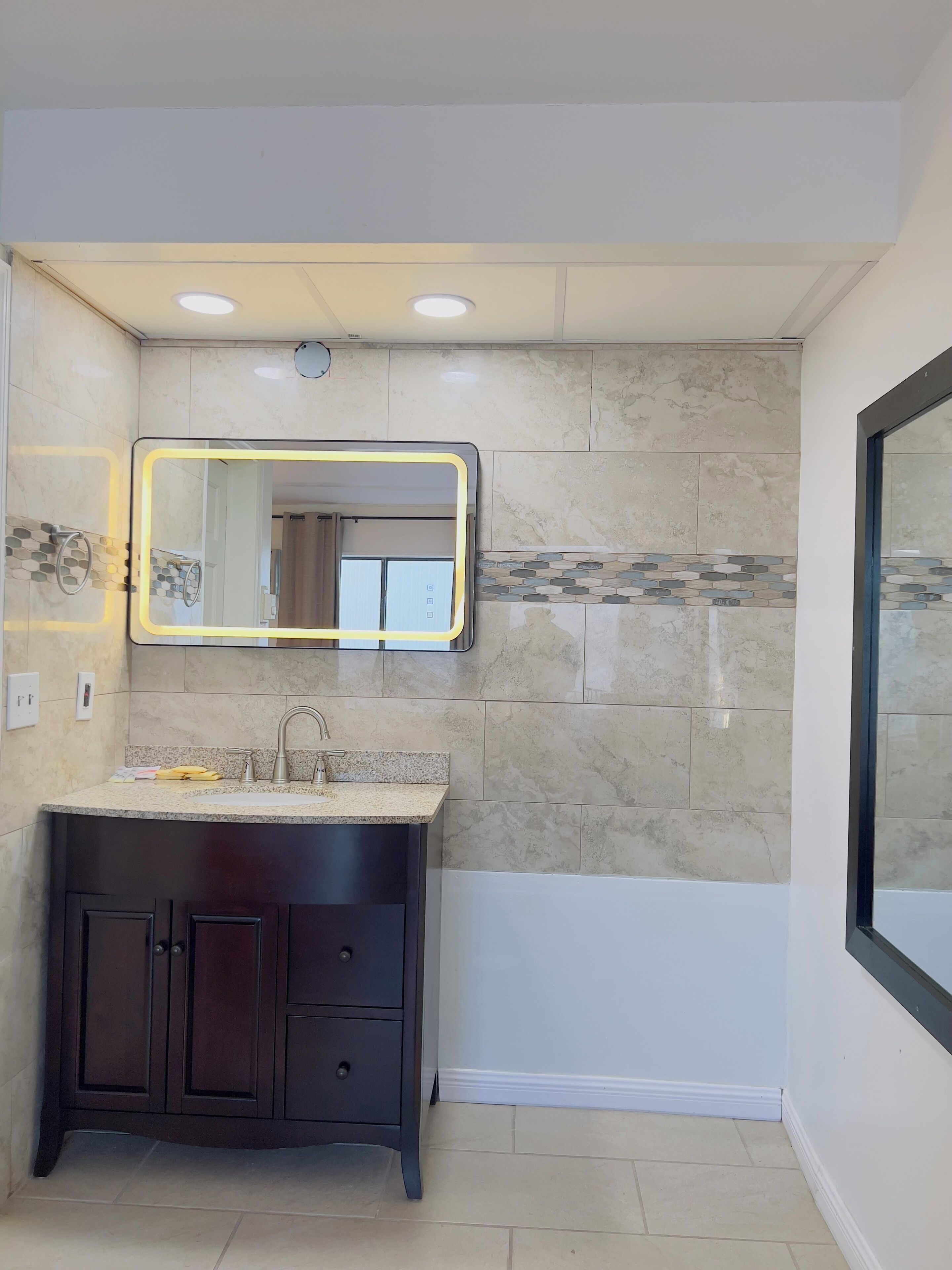 Un baño con tocador de madera oscura, un espejo grande con marco iluminado y salpicadero de azulejos.
