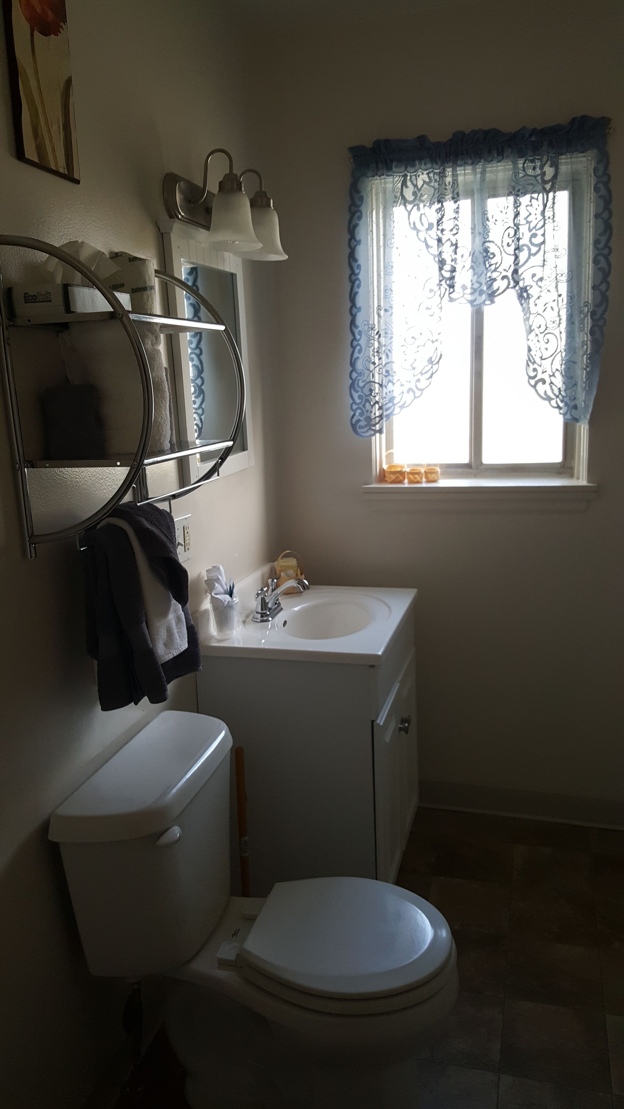 Un bagno con servizi igienici, lavandino e specchio. Si vede una finestra con una tenda blu.