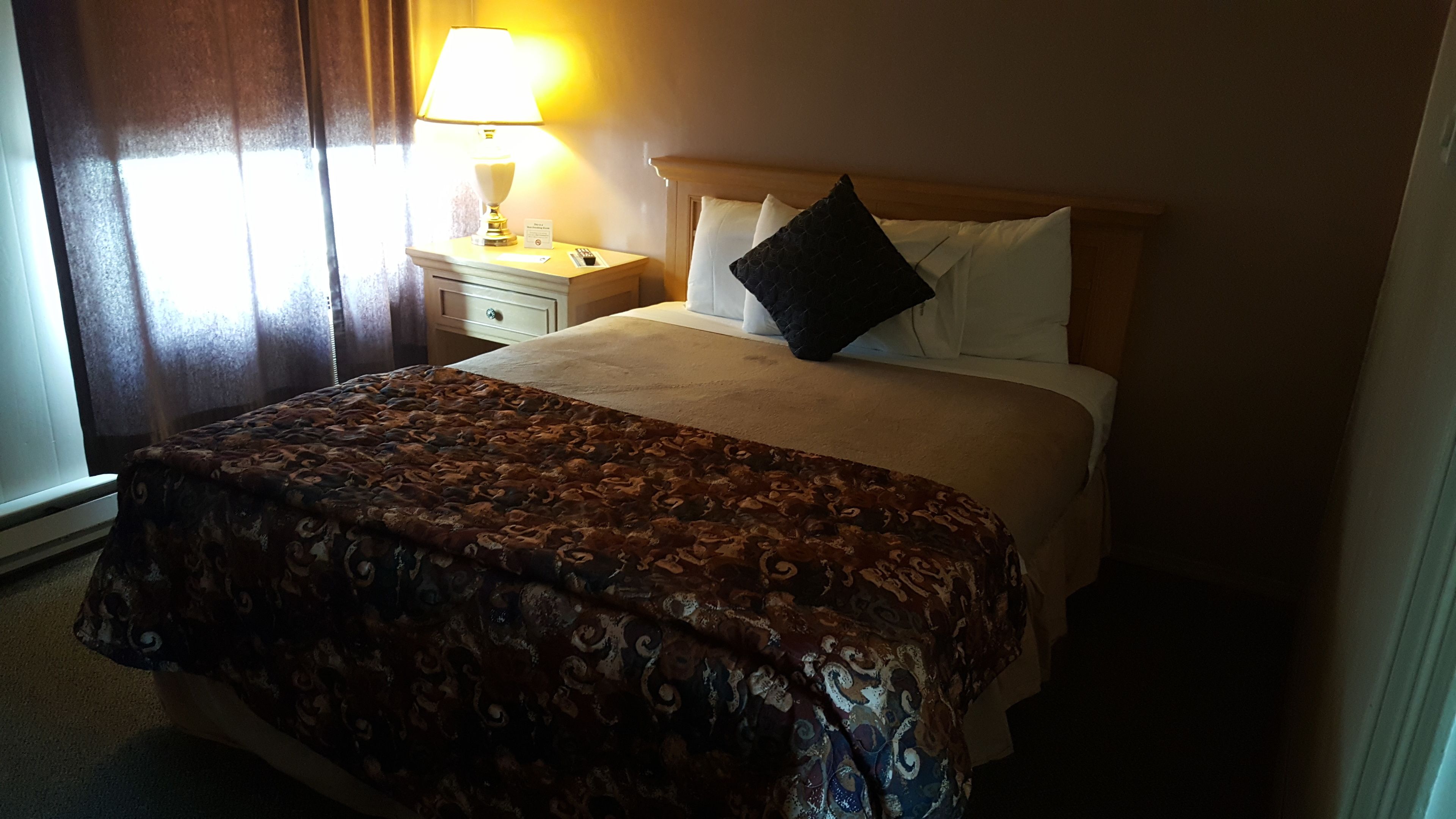 A hotel room with a bed, a bedside table with a lamp, and a window with curtains.