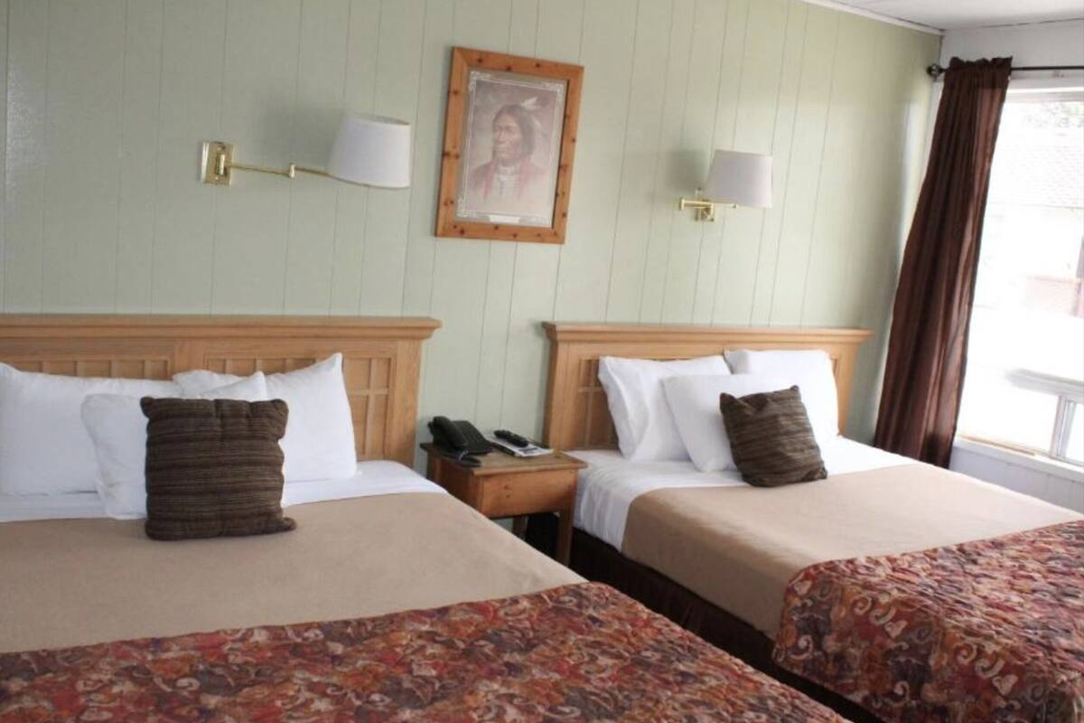A hotel room with two beds, each with a wooden headboard, white bedding, and a patterned throw. There are bedside tables with lamps and a framed picture on the wall.