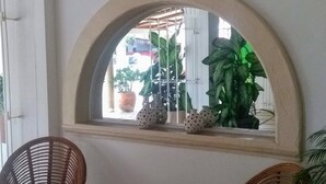 Un área de recepción con un letrero que dice 'WELCOME' sobre una ventana, sillas de mimbre, una mesita redonda y plantas en macetas.