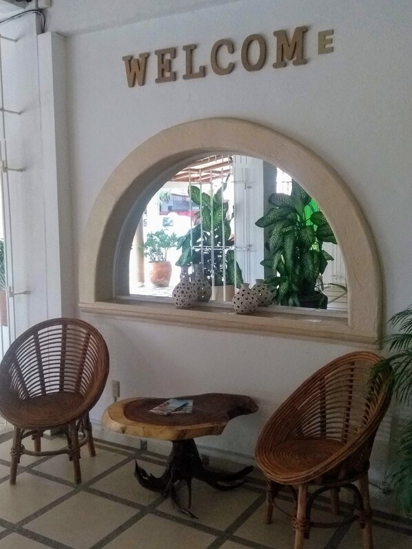 Uma área de recepção com um letreiro 'WELCOME' sobre uma janela, cadeiras de vime, uma pequena mesa redonda e plantas em vasos.