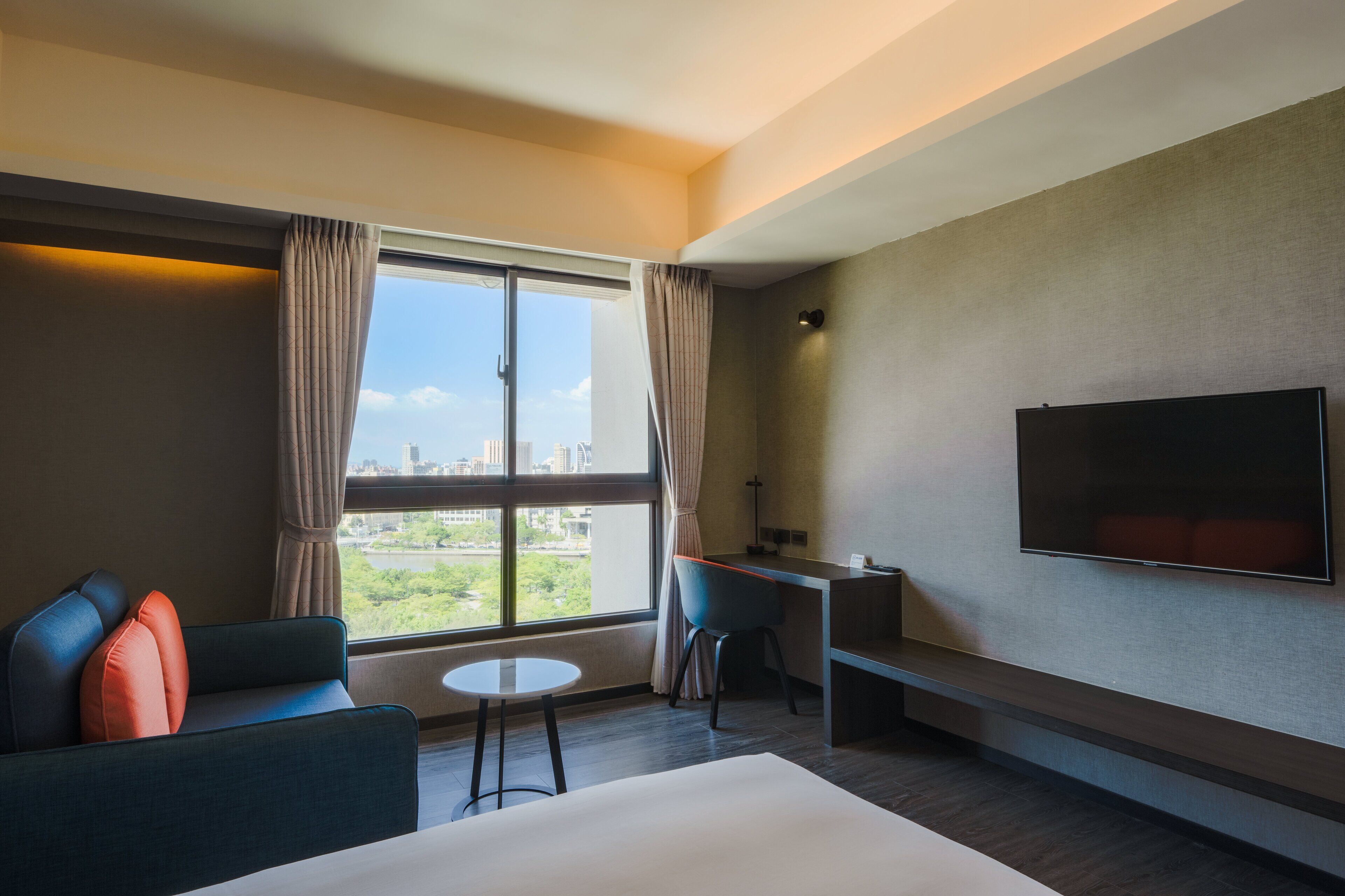 A modern hotel room with a large window, a flat-screen TV, a sofa, a desk, and a small table.