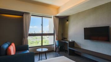 A modern hotel room with a large window, a flat-screen TV, a sofa, a desk, and a small table.
