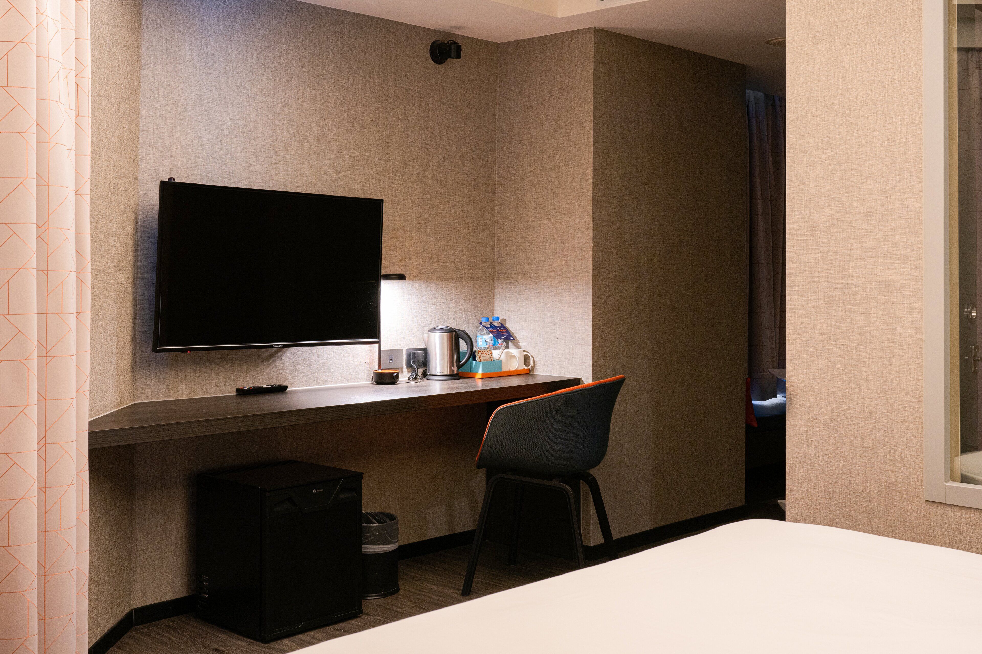 Quarto de hotel com televisão de tela plana na parede, pequena escrivaninha com cafeteira, cadeira e lixeira.