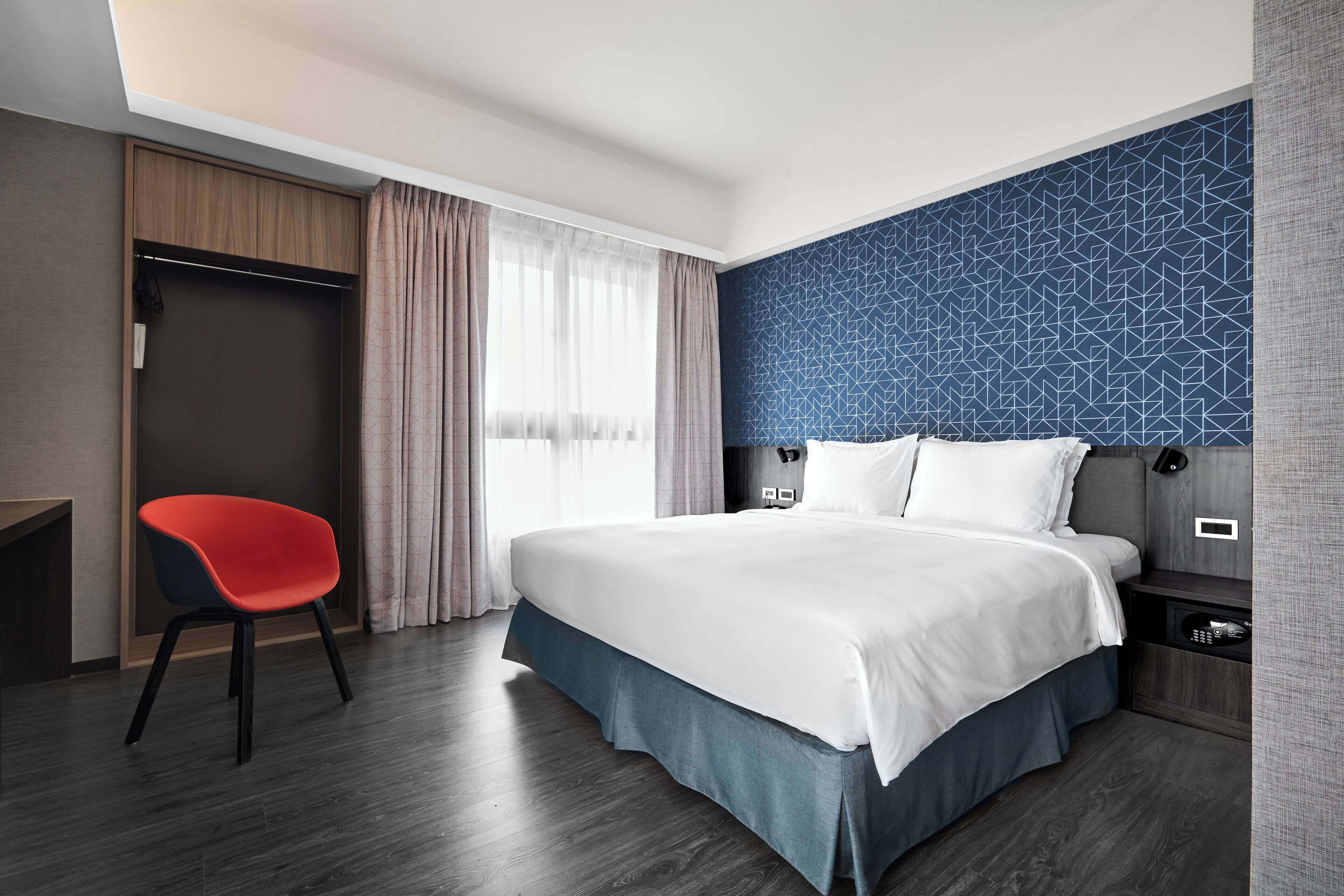 Quarto de hotel com cama, cadeira, guarda-roupa e parede com padrão geométrico.