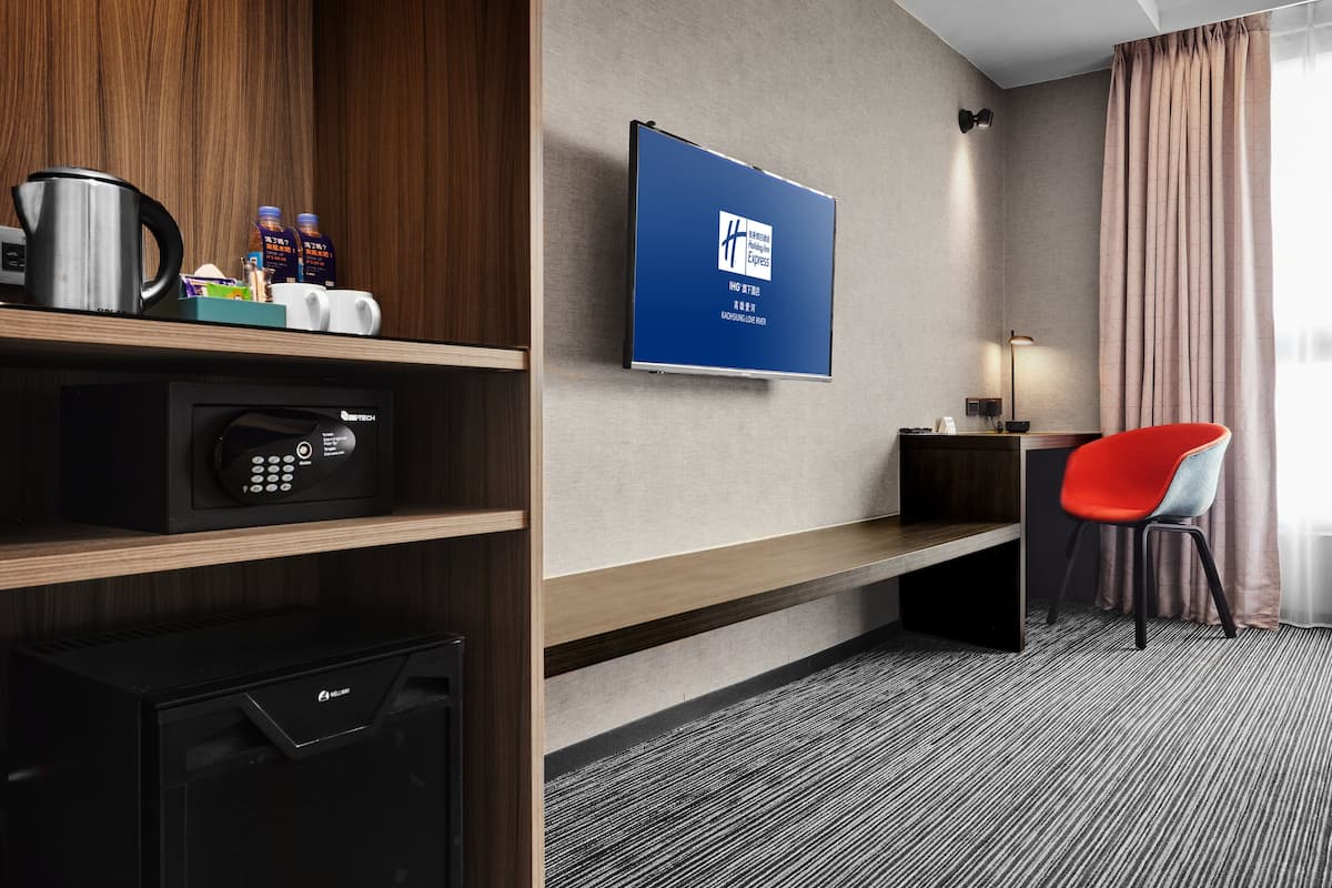Habitación de hotel con microondas, un refrigerador pequeño, cafetera y un televisor de pantalla plana montado en la pared. Hay una silla roja y una mesita con una lámpara.