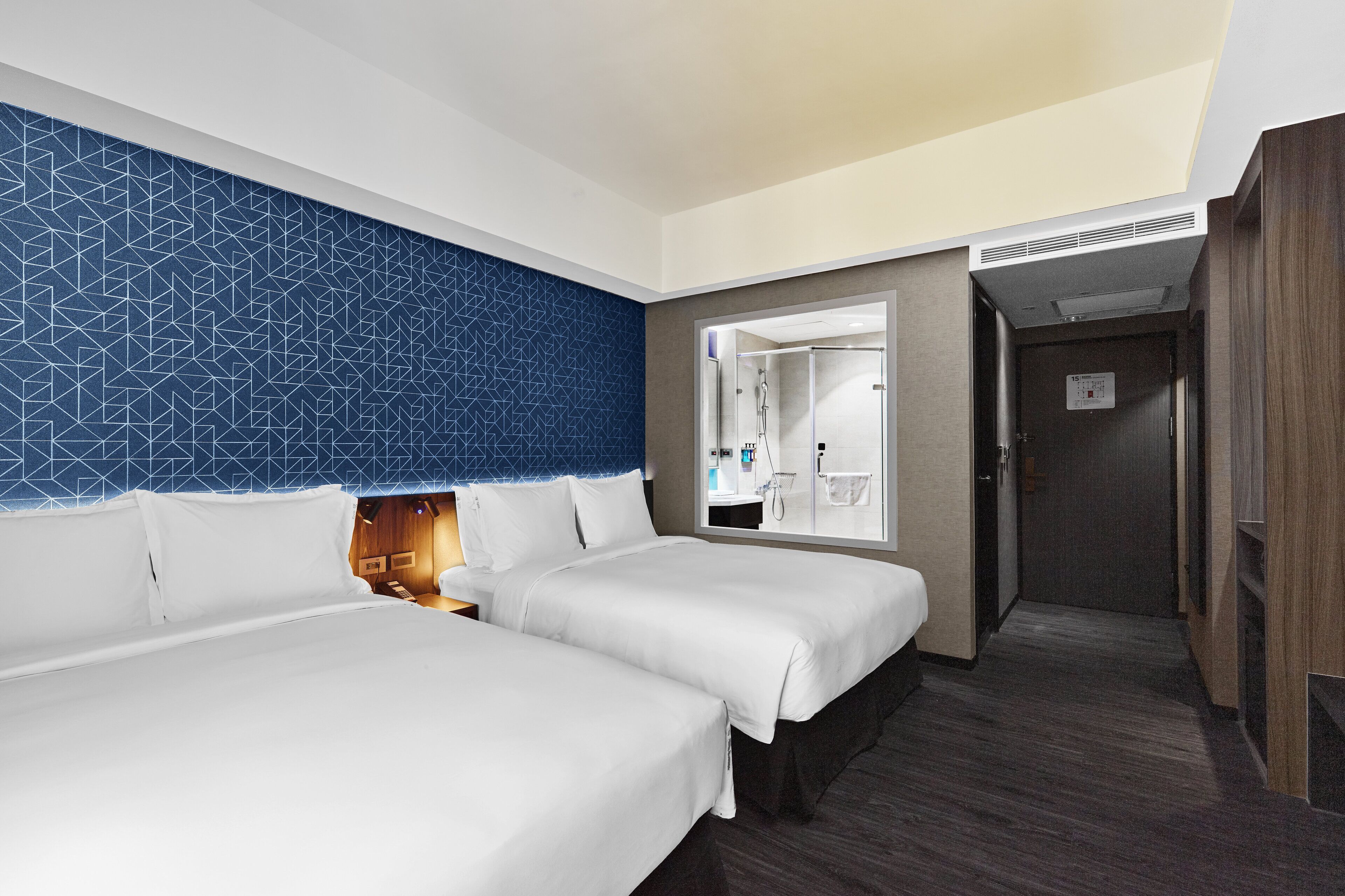 Quarto de hotel com duas camas, parede azul com padrão geométrico, um espelho grande e área de chuveiro.
