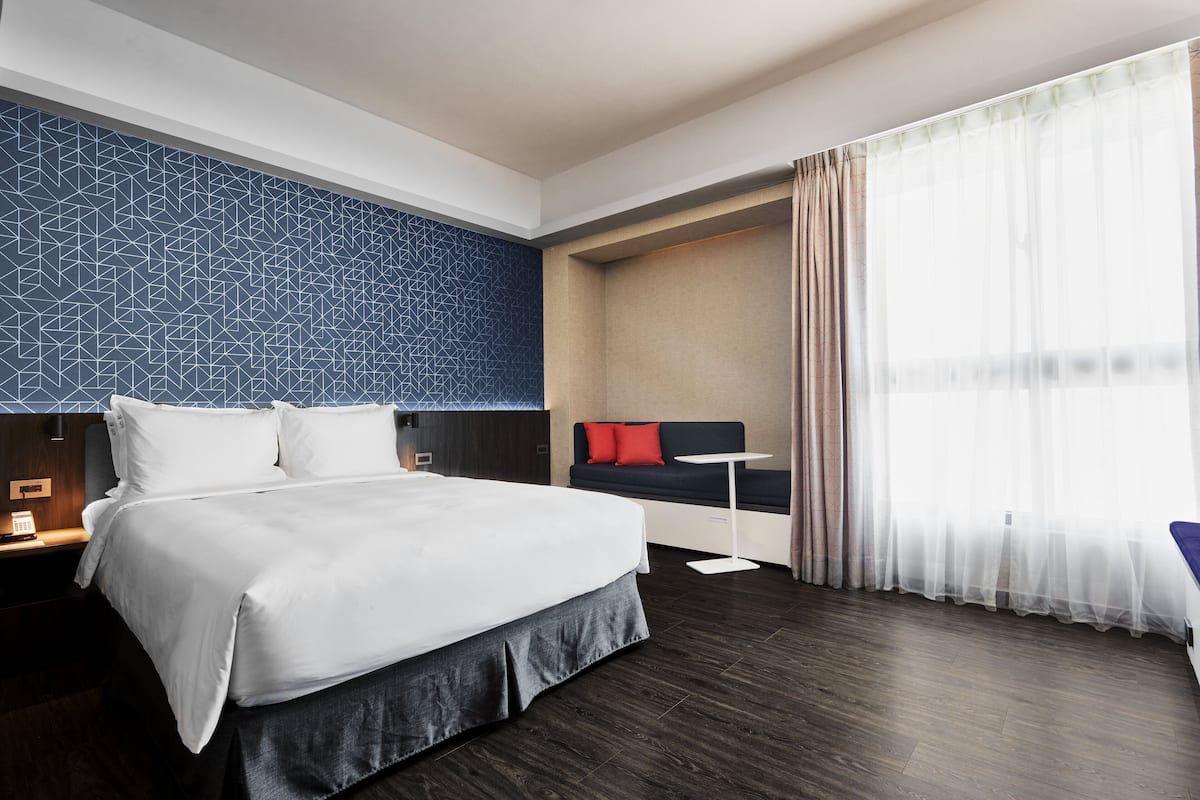 Una habitación de hotel moderna con una cama grande, un área de descanso con sofá y una mesita, y un ventanal con cortinas translúcidas.