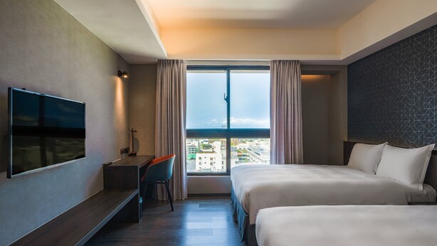 A hotel room with two beds, a TV, a desk, and a large window with a city view.