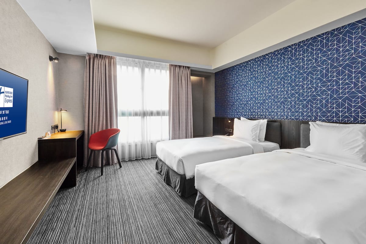 Habitación de hotel con dos camas, un escritorio, una silla y un televisor en la pared.