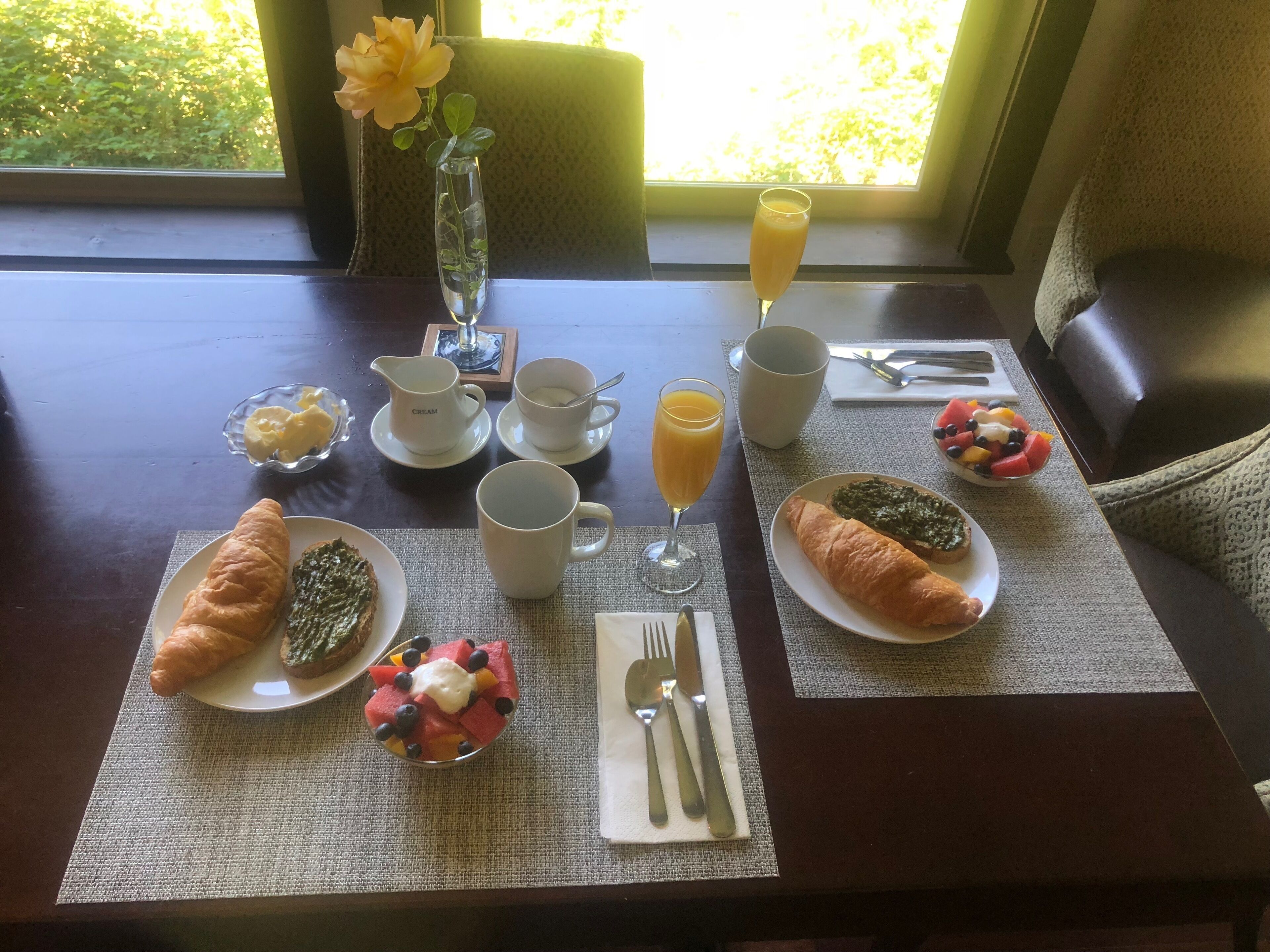 A breakfast table with croissants, fruit, juice, and a vase with a flower.