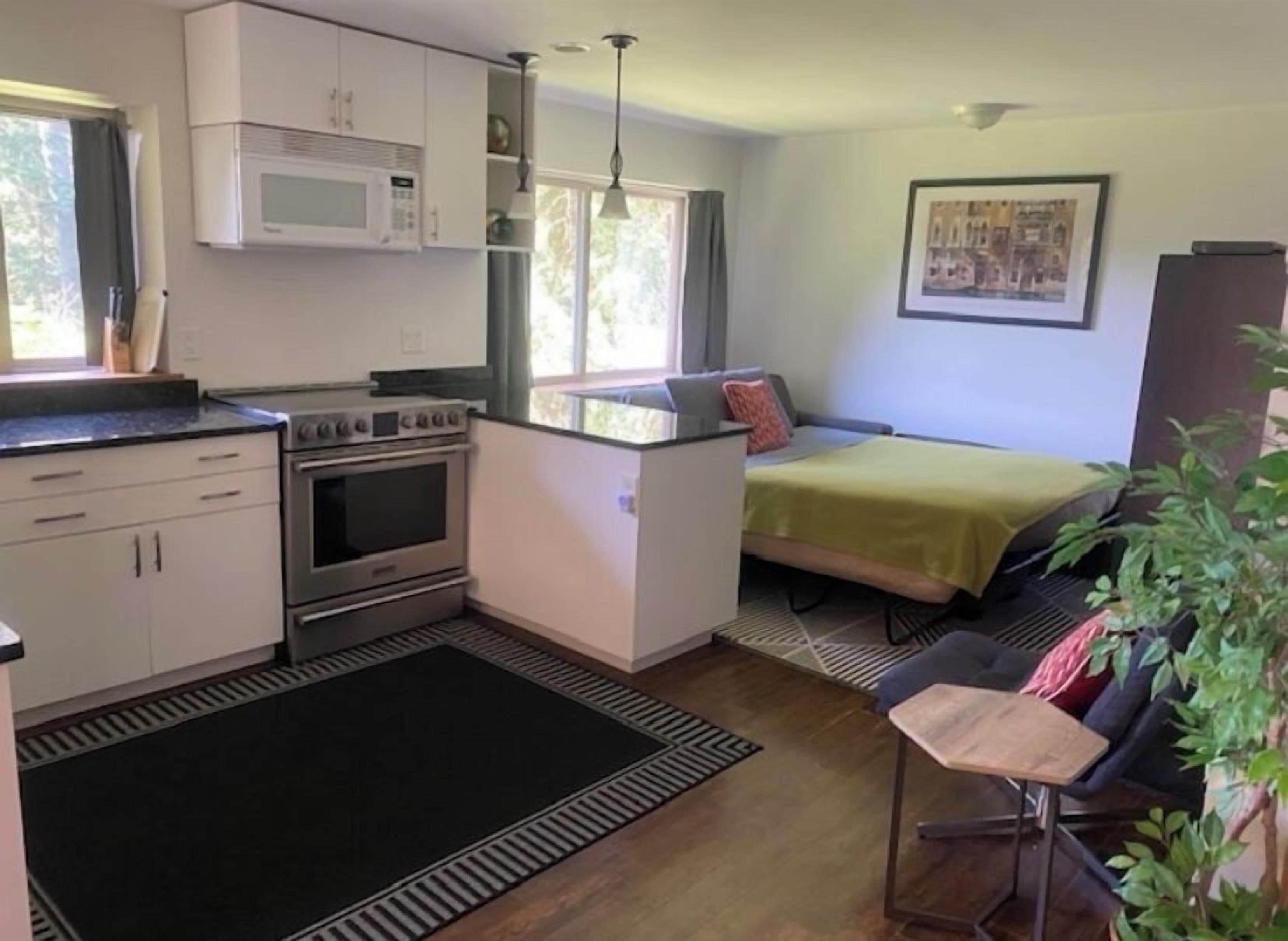 Cocina compacta con microondas, horno y estufa, un pequeño comedor con mesa y sillas, y una cama con manta verde.