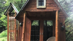 A wooden cabin named 'Twin Cedars Chapel' with a porch, a wicker chair, and a small statue. - Davis Brook Retreat (Sechelt)