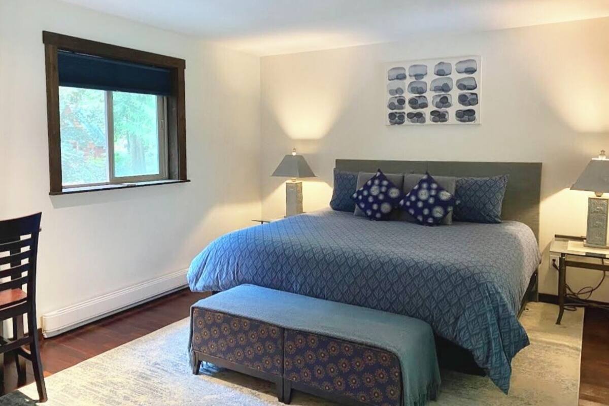 A bedroom with a bed, two bedside lamps, a window, a chair, and a wall with a picture.