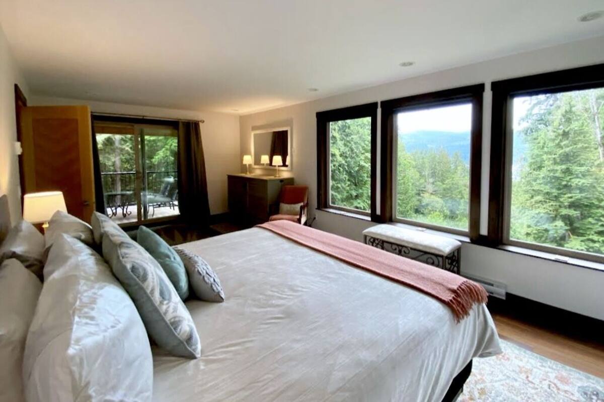 A bedroom with a large bed, a desk, and a view of the forest through large windows.