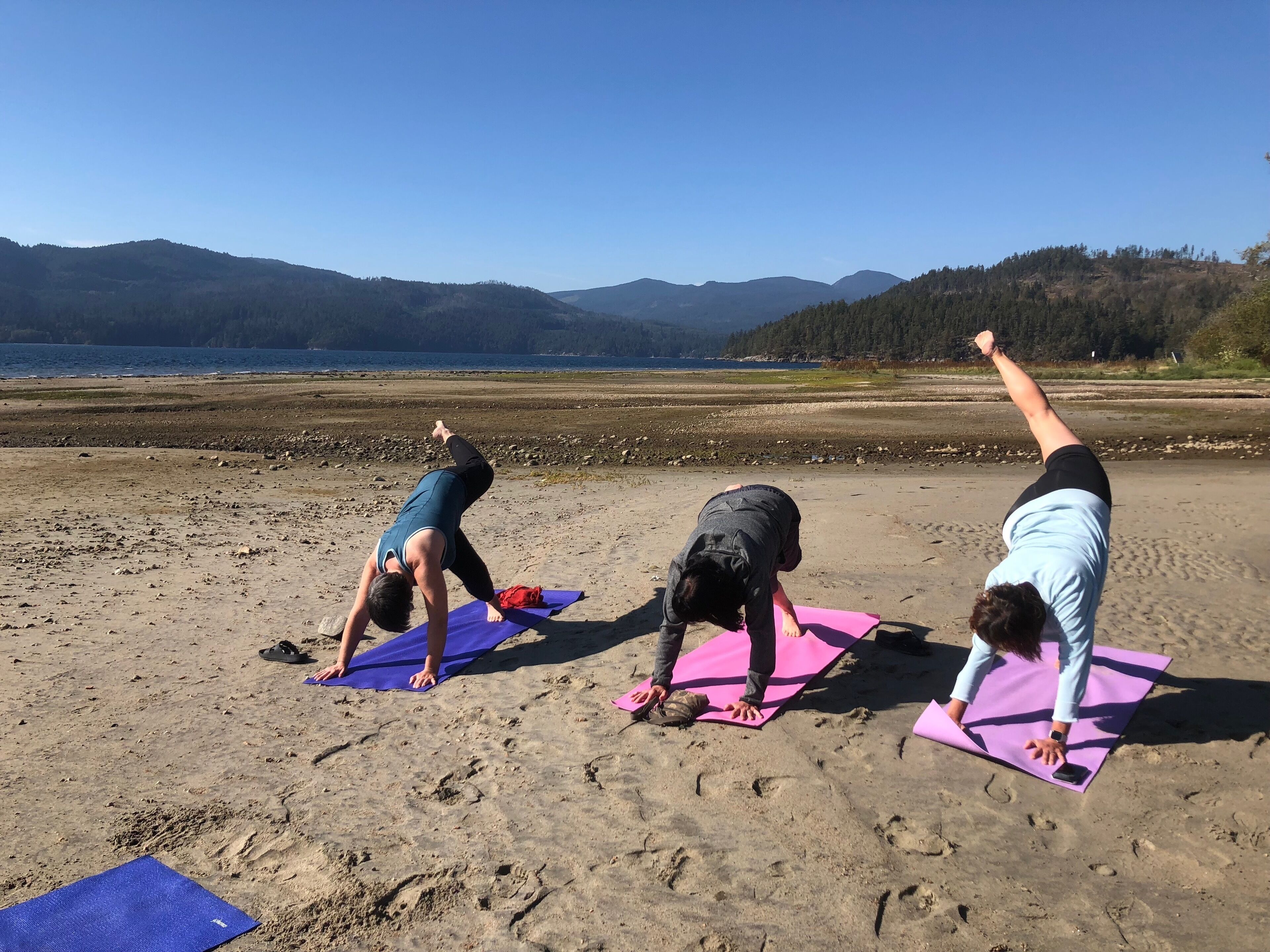 Three people performing yoga on mats on a beach with mountains in the background.