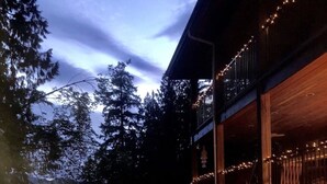 A hot tub in a backyard with a wooden deck and a building with string lights. - Davis Brook Retreat (Sechelt)