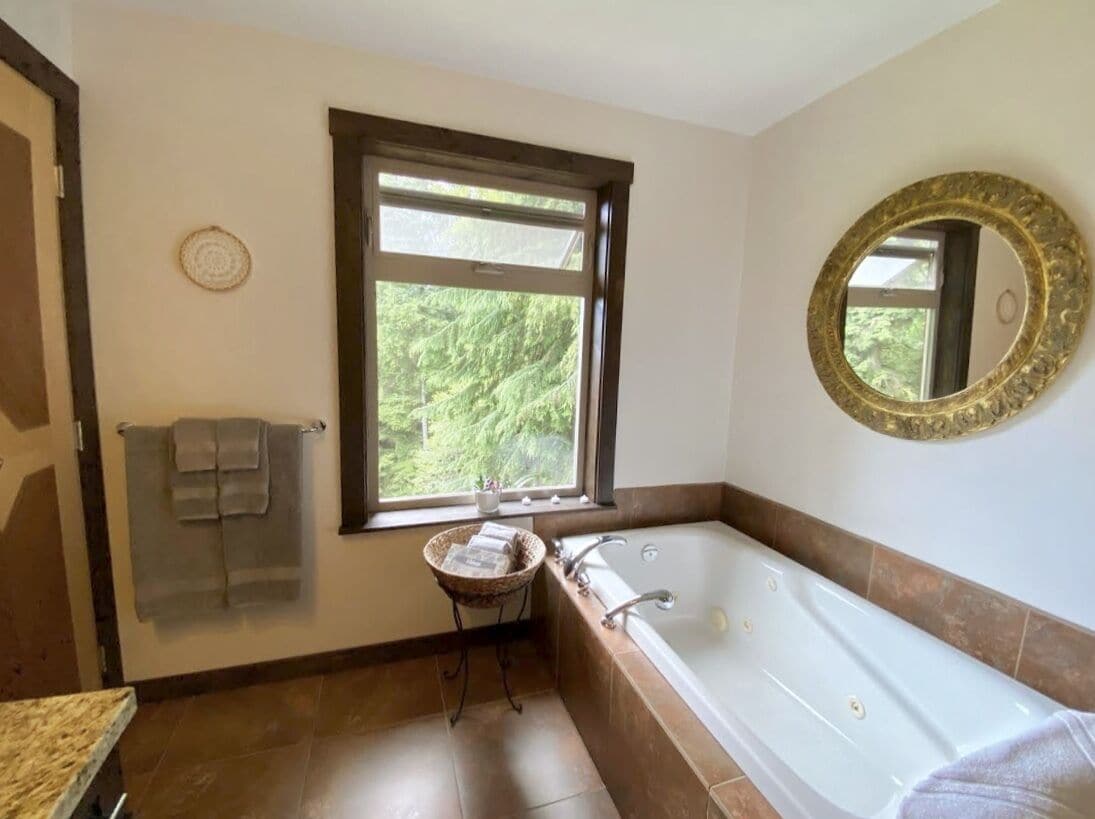 Un baño con una bañera grande, una ventana con vistas a la vegetación, un perchero y un espejo decorativo.