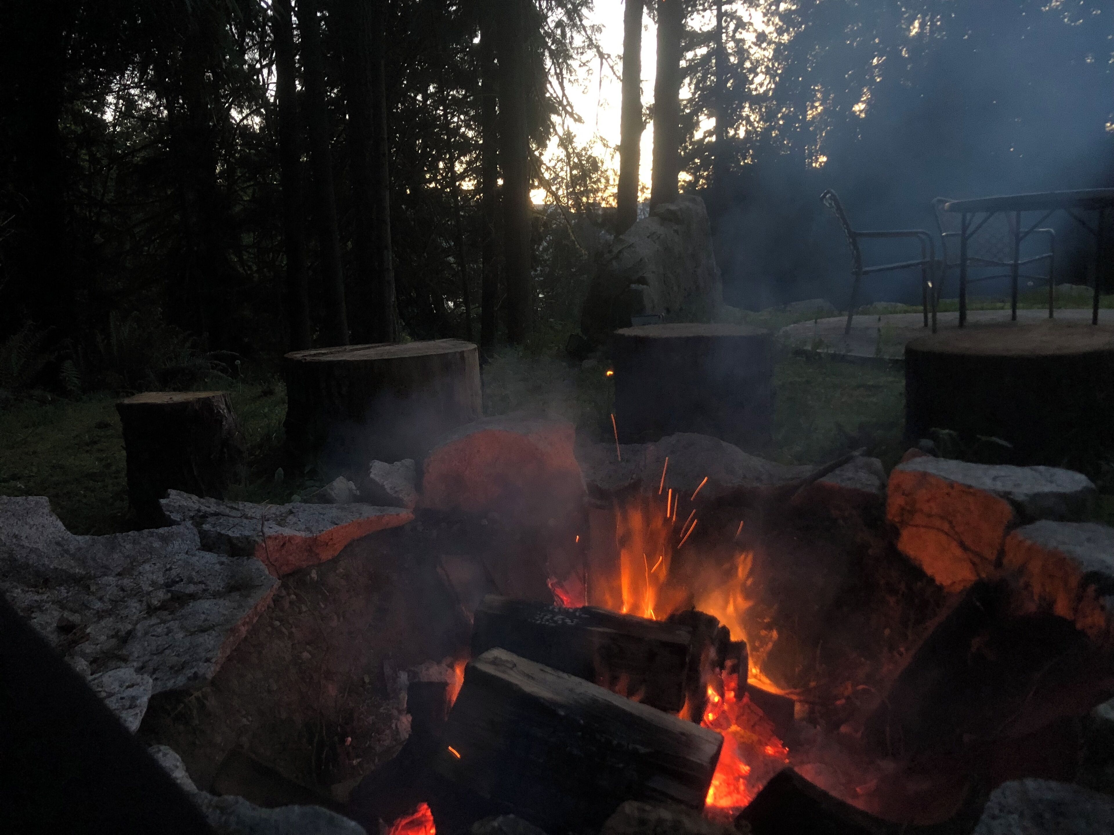 A campfire surrounded by rocks and logs, with a chair and table nearby.