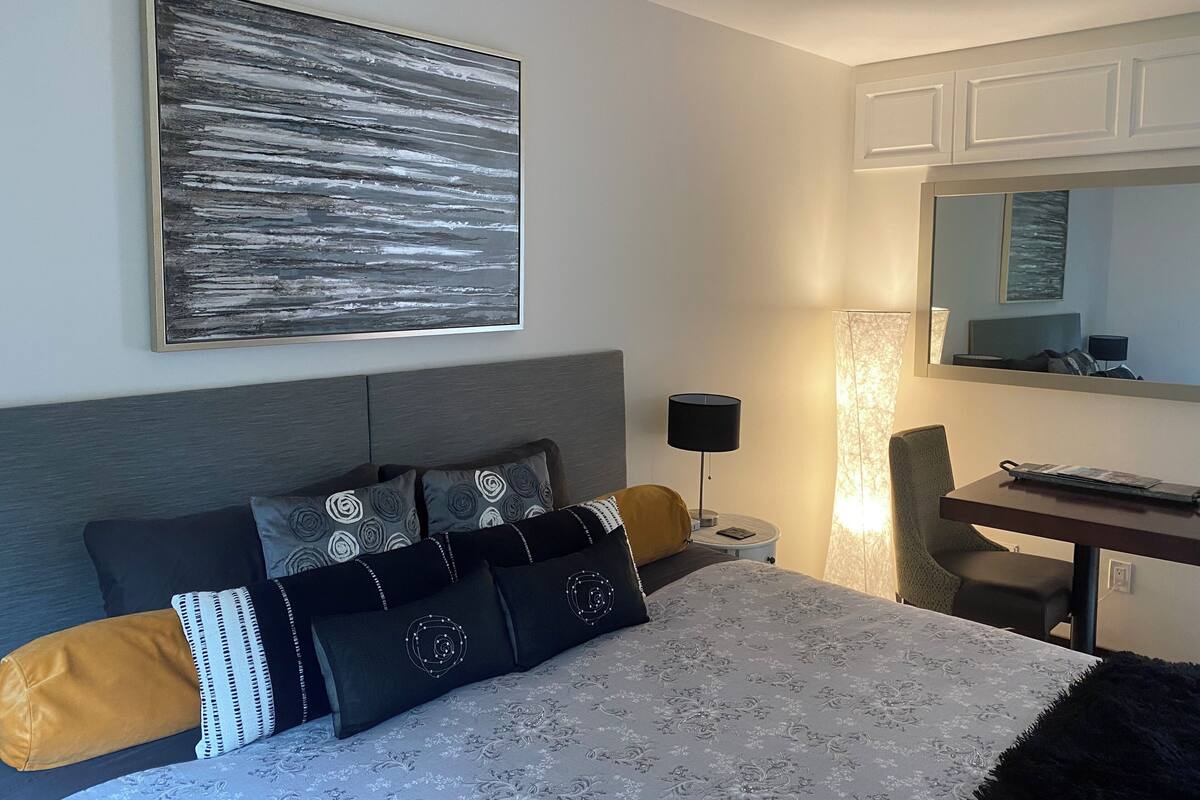 A modern bedroom with a large bed, a desk, and a chair. There is a painting on the wall and a lamp on the desk.
