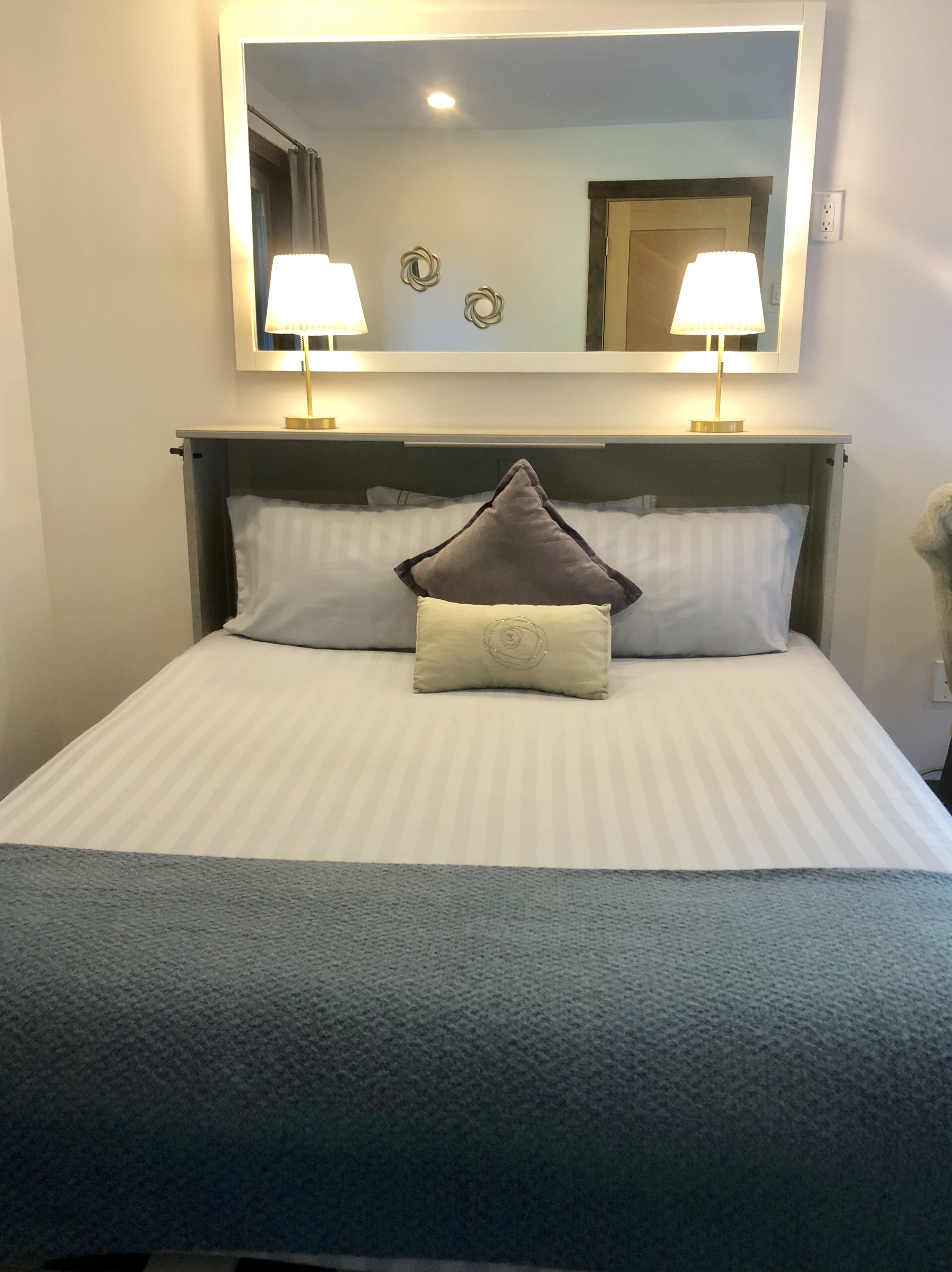 A neatly made bed with a grey and white striped bedspread, two bedside lamps, and a large mirror above the bed.