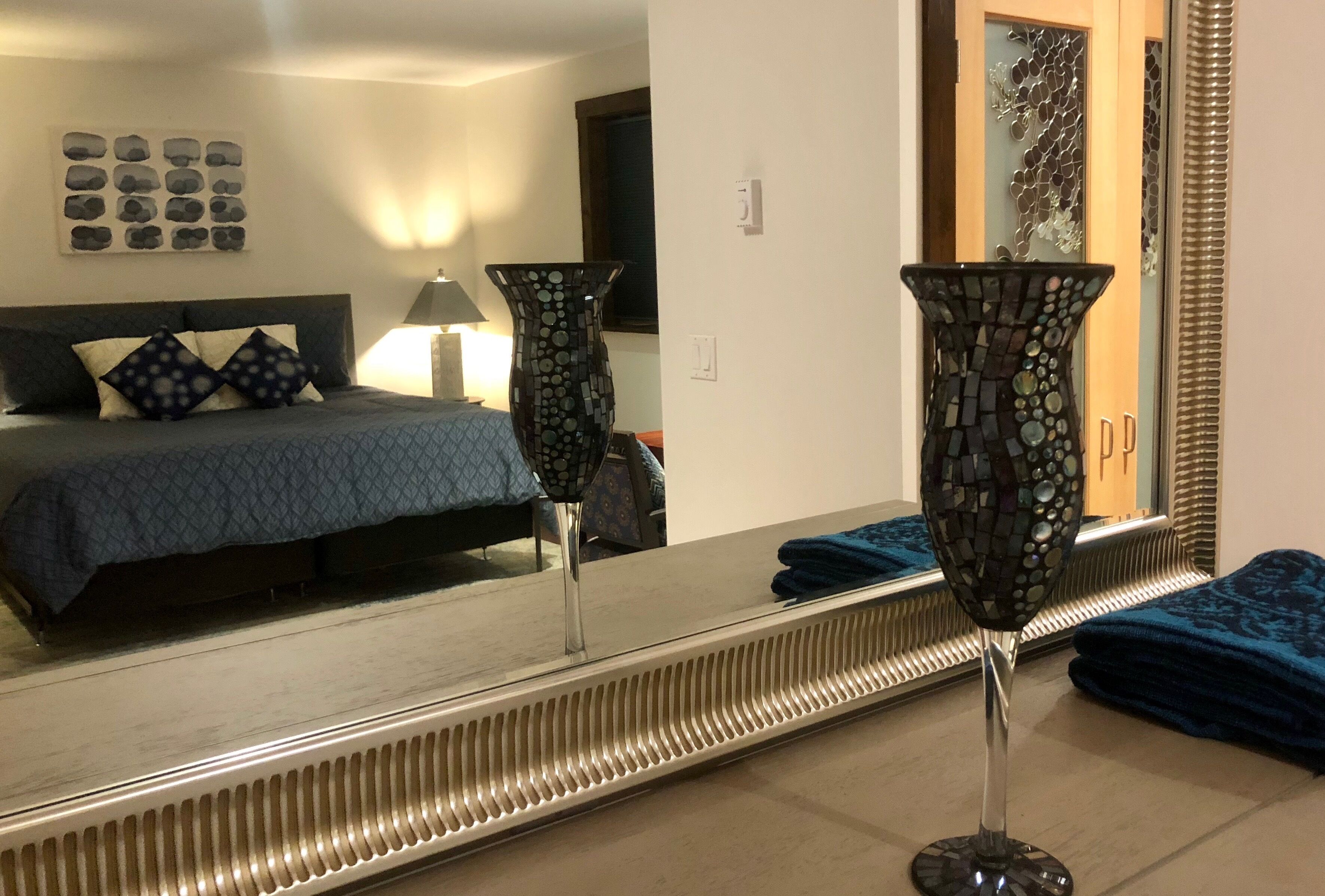 Habitación de hotel con una cama, un espejo grande, un jarrón decorativo y un baño con espejo y lavamanos.