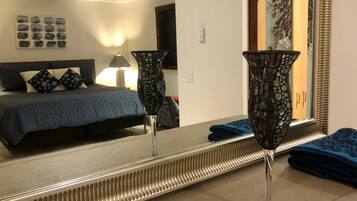 Habitación de hotel con una cama, un espejo grande, un jarrón decorativo y un baño con espejo y lavamanos.