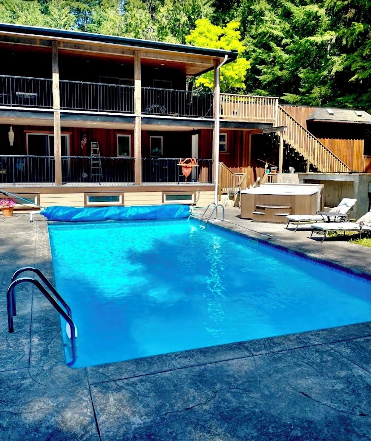 A swimming pool with a wooden deck and lounge chairs, surrounded by a building with a balcony and a staircase.