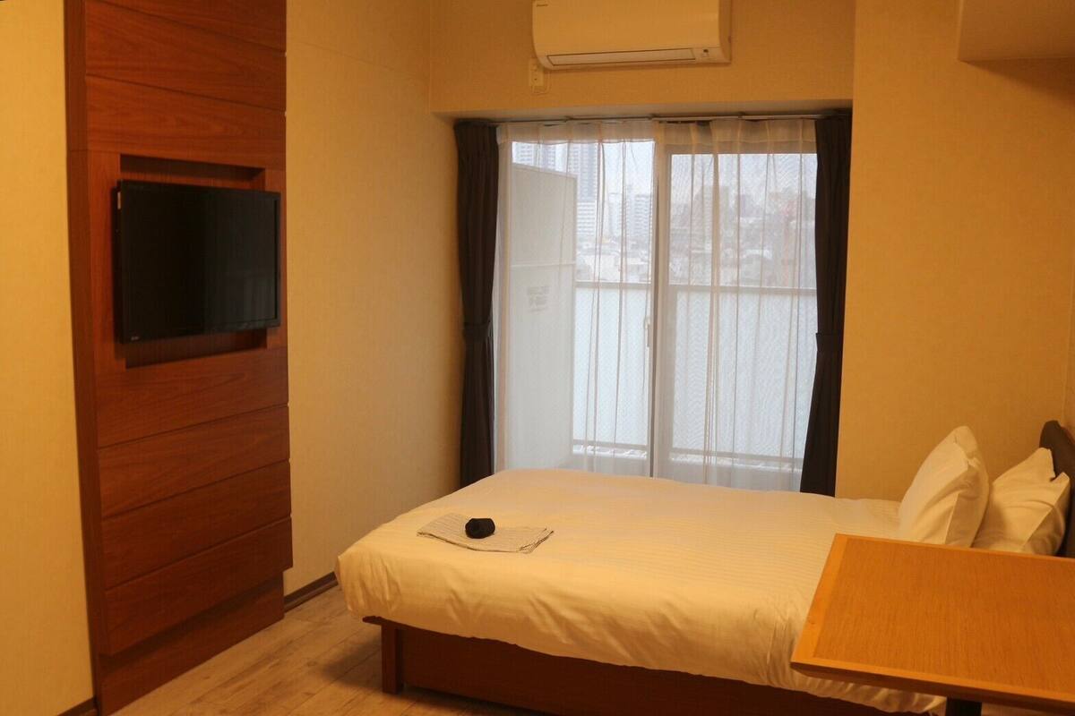 Une chambre à coucher avec un lit, une armoire en bois, un téléviseur fixé au mur et une fenêtre avec des rideaux.