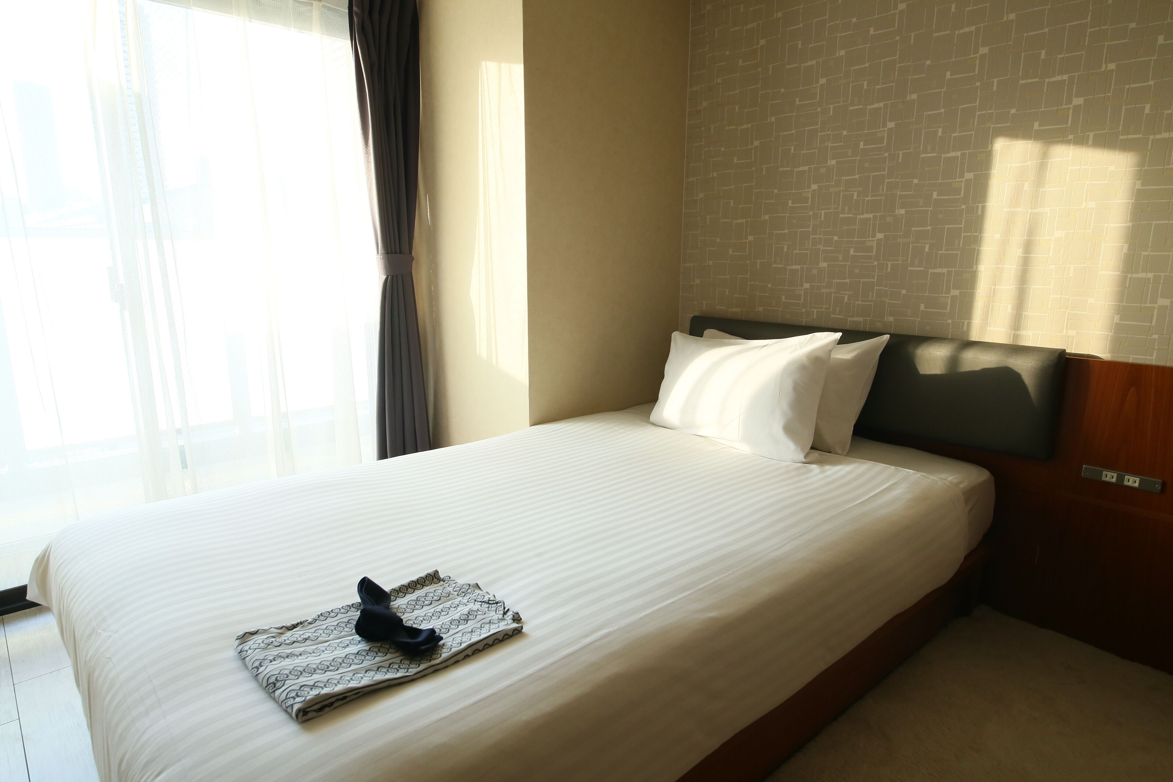 A neatly made bed with white linens and a patterned pillow, a wooden headboard, and a brick-patterned wall.